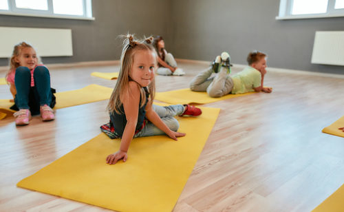Portrait of a cute little girl sitting on yellow yoga mat and looking at camera with smile. Group of children lying on yoga mats and relaxing after gymnastic class. Dance school. Taking a break